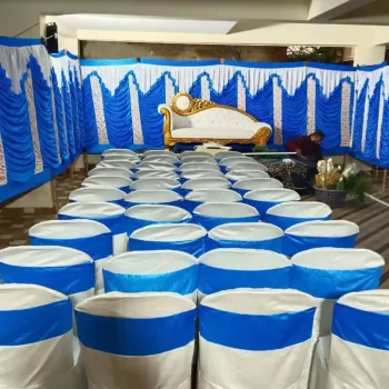 Tent setup by HKGN Tent House & Decoration in Hyderabad, showing white chairs dressed in blue and white fabric and a plush sofa set against blue and white drapes, giving the space a warm, festive feel.