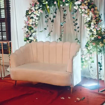 Floral setup by HKGN Tent House & Decoration in Hyderabad: a white loveseat before an arch of white and pink blooms and greenery, set on a red carpet.